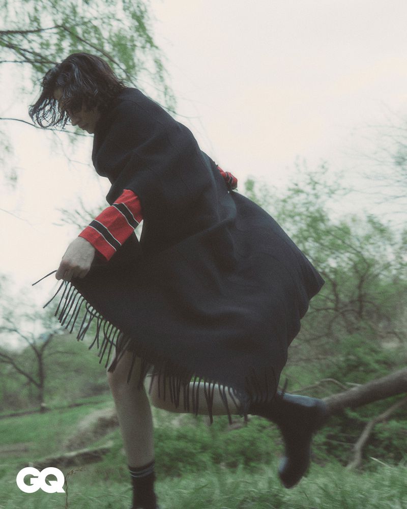 Fashion editorial shot of a woman in a black poncho with fringe walking in a grassy field with trees and branches in the background.