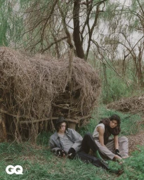 Two male models seated in a woodland setting next to a large woven brush structure for a GQ Korea editorial.
