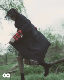 Model in black fringed poncho and red striped sleeves running through a grassy outdoor woodland setting.