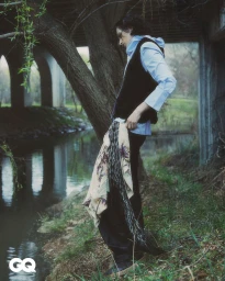 Model standing outdoors near a riverbank under a concrete bridge wearing a light blue shirt and dark vest for GQ Korea.