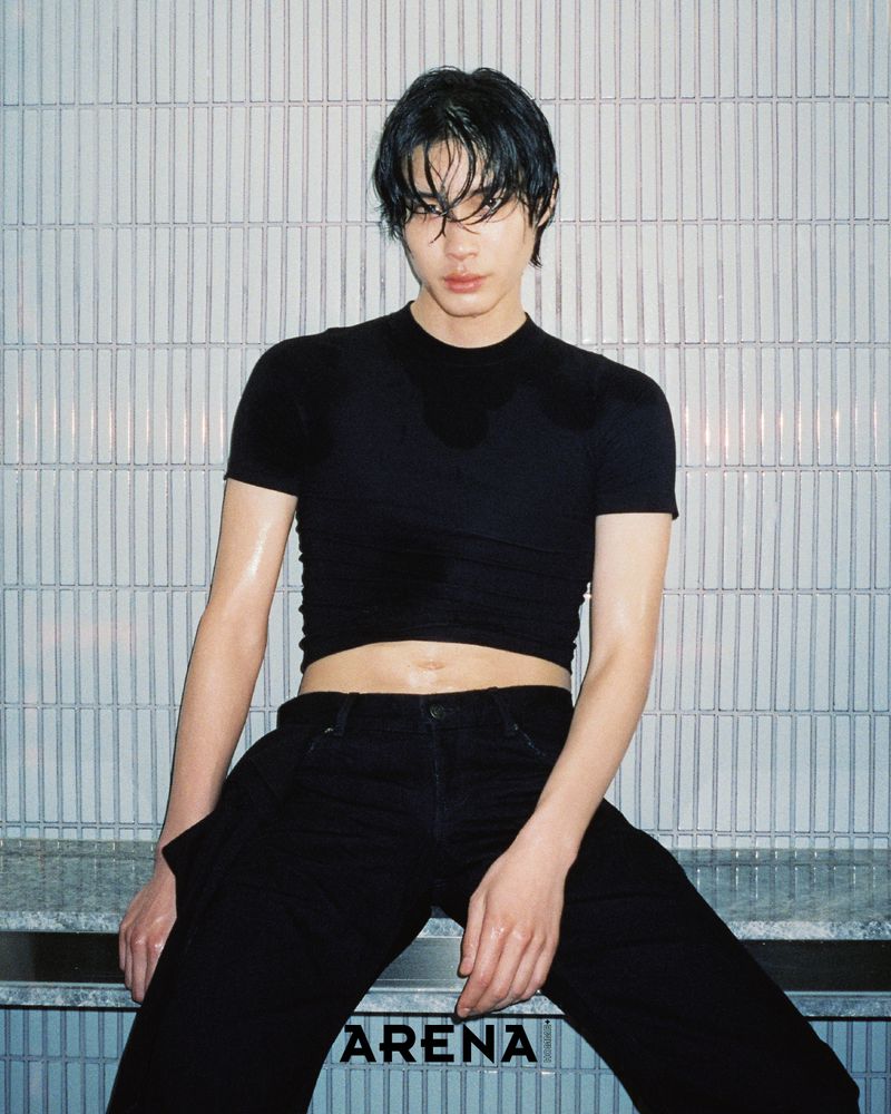 Portrait of a young adult male with dark hair, wearing a black top and jeans, seated against a textured white wall with visible 'Arena' text.