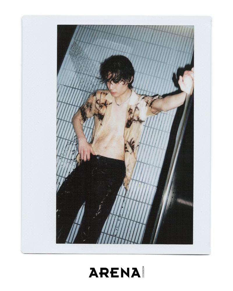 Portrait of a wet, shirtless adult male in a tropical shirt and dark pants, posed in front of a metallic striped wall with Arena branding.