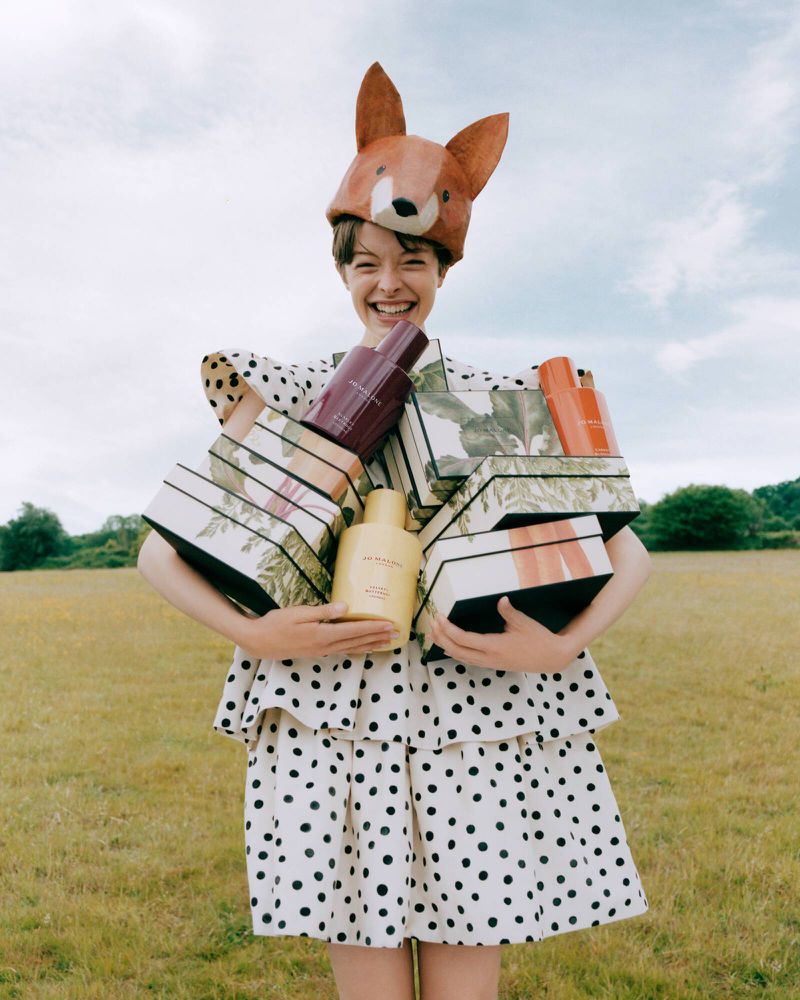 Model wearing a fox hat and polka dot dress holding stacked Jo Malone fragrance boxes in a meadow