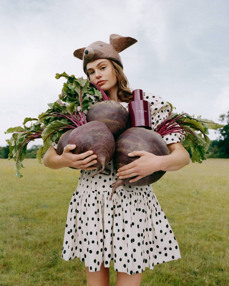 Model wearing a mouse ear hat and polka dot dress holding giant beetroots and a maroon perfume bottle in an open grassy field.
