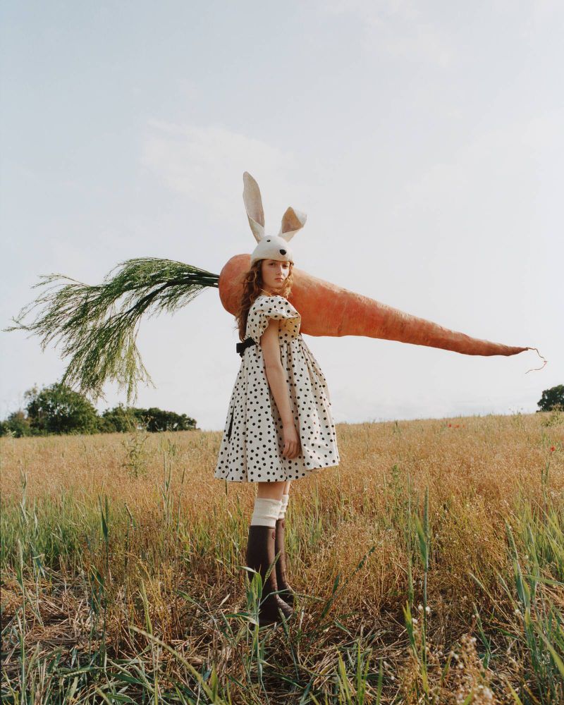 Model wearing a polka dot dress and bunny headpiece balancing a giant oversized carrot on her shoulder in an open grassy field.