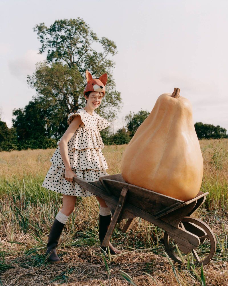 Model in polka dot dress and animal ears pushing a wheelbarrow with a giant gourd in a grassy field photographed by Toby Coulson.