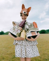 Model wearing a fox hat and polka dot dress holding stacked Jo Malone fragrance boxes in a meadow