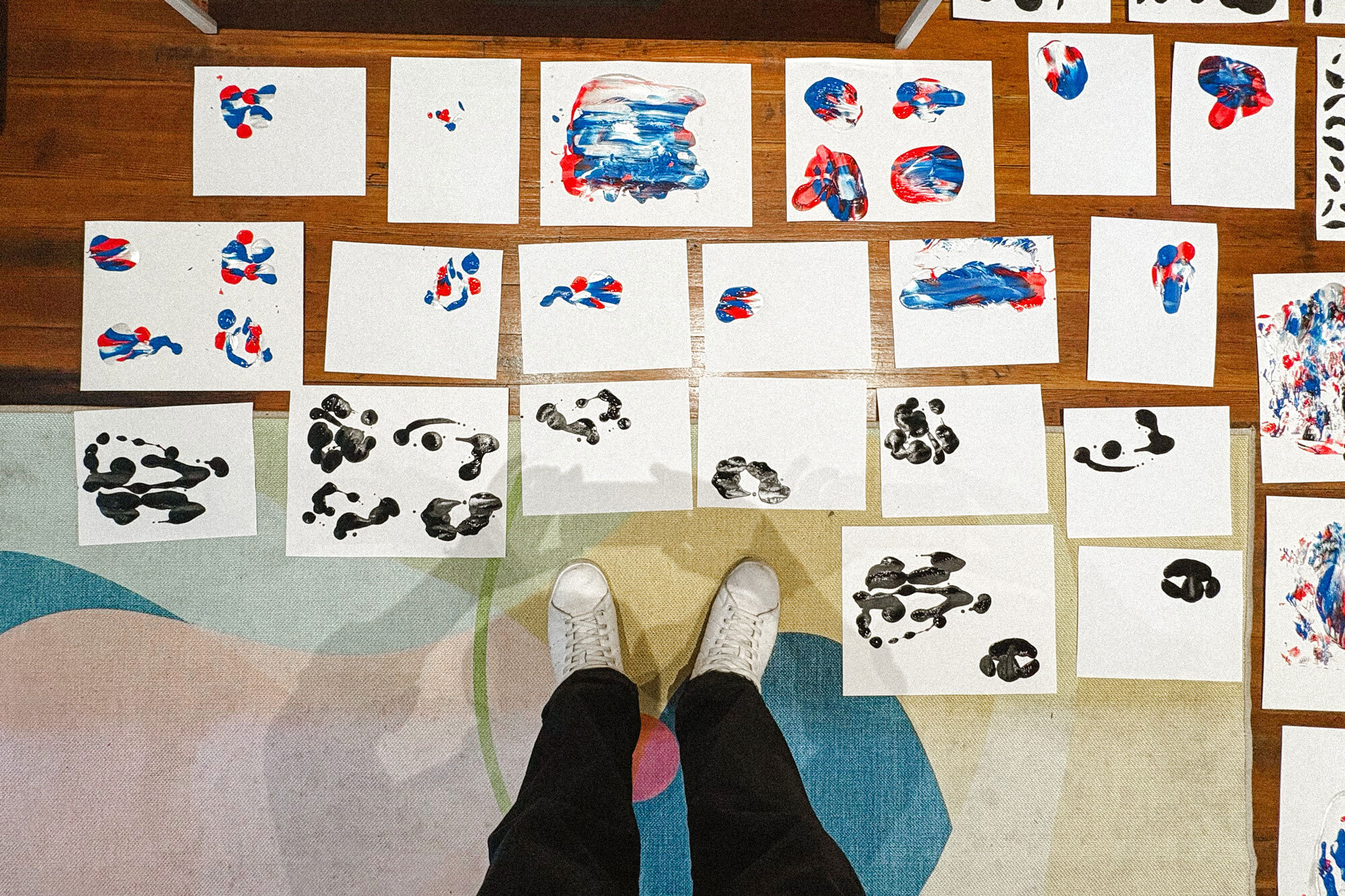 Overhead point-of-view shot of white sneakers standing near paper sheets with red, blue, and black paint prints on a wood floor.