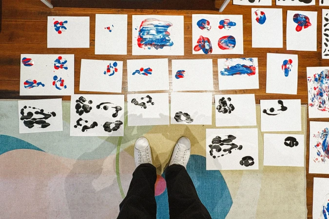Overhead point-of-view shot of white sneakers standing near paper sheets with red, blue, and black paint prints on a wood floor.