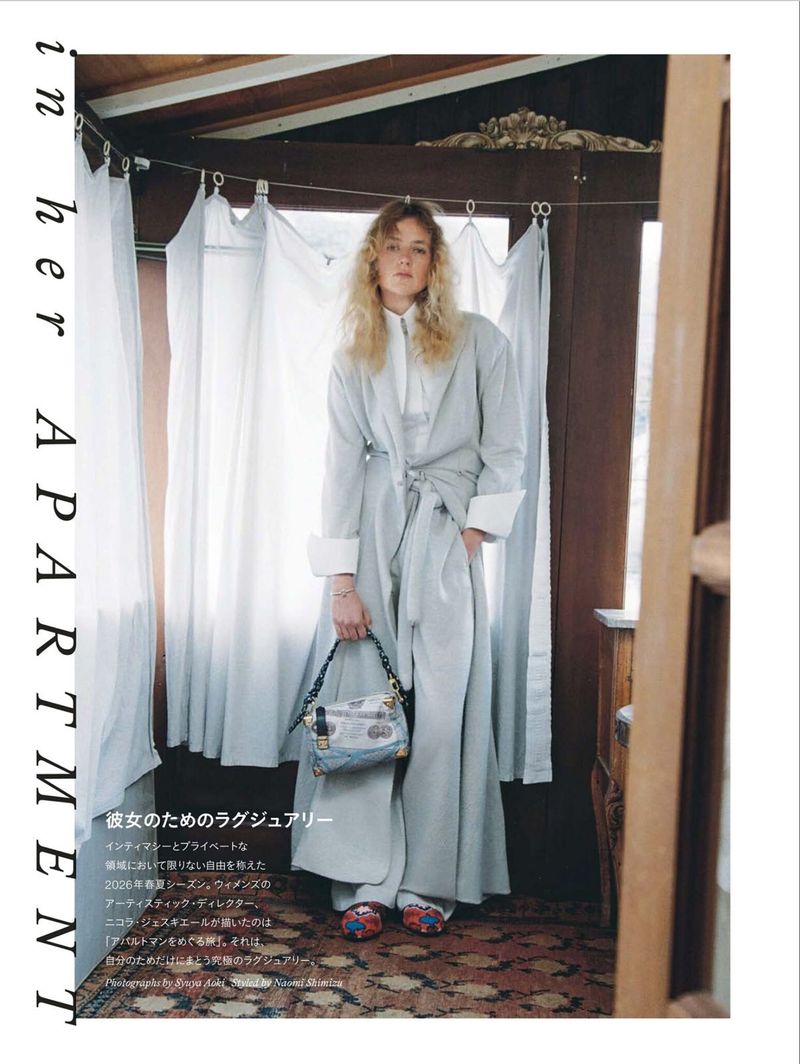 Model Thialda Bok standing in a light grey robe coat and trousers holding a printed handbag against curtained windows on a patterned rug.