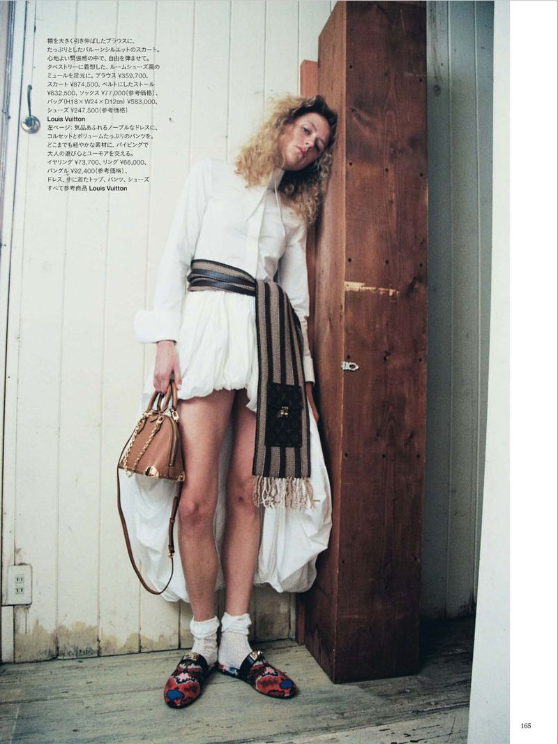Model with curly blonde hair wearing a white dress and patterned scarf leaning against a wooden column in an apartment.