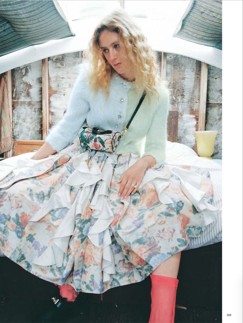 Model with long blonde curly hair wearing a pastel cardigan and floral ruffled skirt seated on a bed in a rustic room.