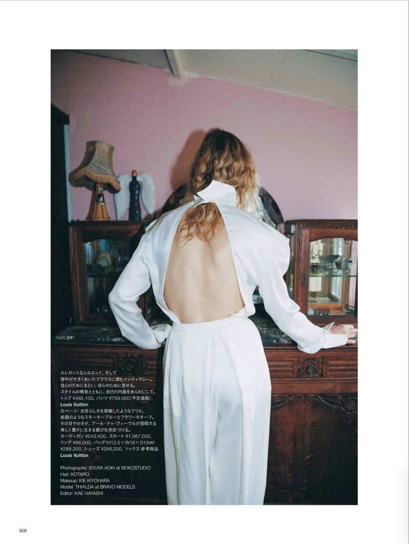 Model Thialda Bok wearing an open-back white blouse standing in a room with pink walls and wooden furniture.