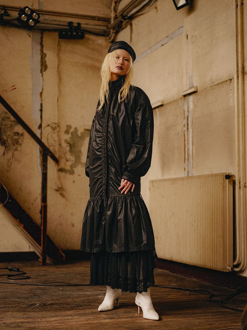Model Seng I U wearing a long black ruched coat dress, black beret, and pointed white boots in a rustic interior setting.