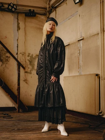 Model Seng I U wearing a long black ruched coat dress, black beret, and pointed white boots in a rustic interior setting.
