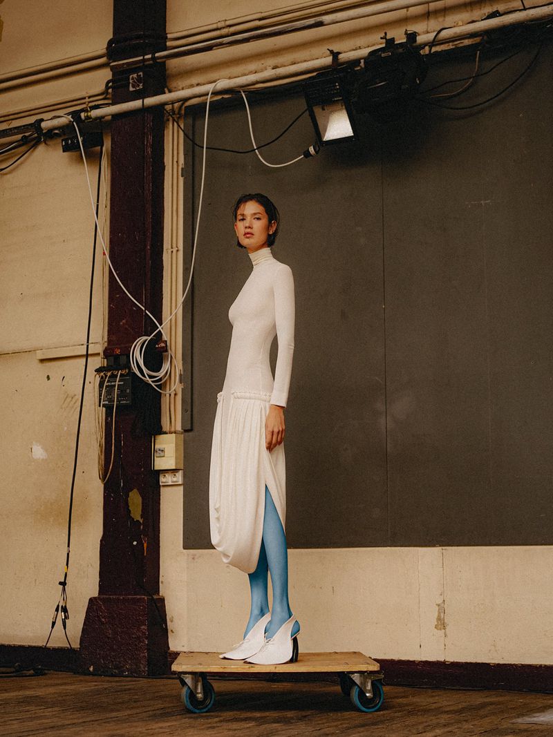 Model standing on a wooden dolly with blue wheels, wearing a long-sleeve white draped dress, vibrant blue tights, and white heels.