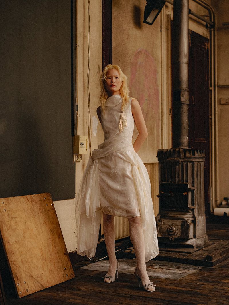 Model posing in a draped off-white sleeveless dress and heels beside a vintage stove in a rustic studio interior.