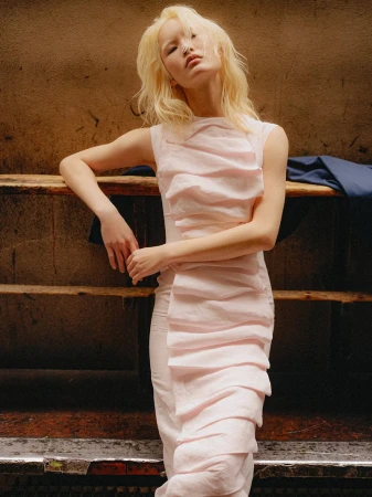 Model with platinum blonde hair wearing a pale pink ruched sleeveless dress leaning against a wooden barrier.