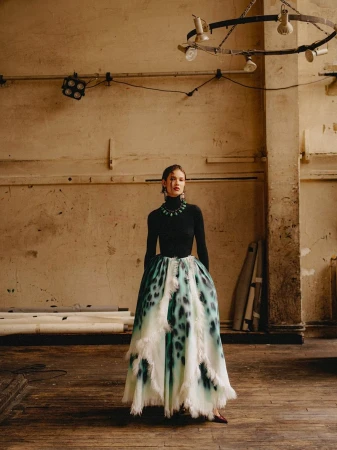 Fashion model in a black turtleneck top and a voluminous tiered white and teal skirt standing on a wooden floor in a studio.