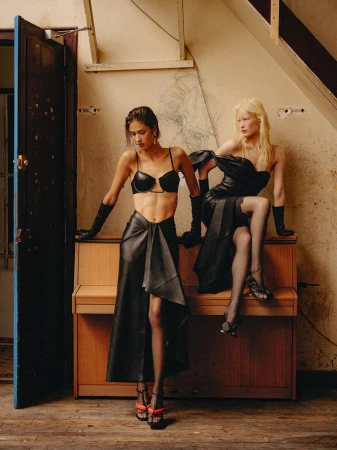Two fashion models in black eveningwear and opera gloves posing beside and seated on an upright wooden piano.