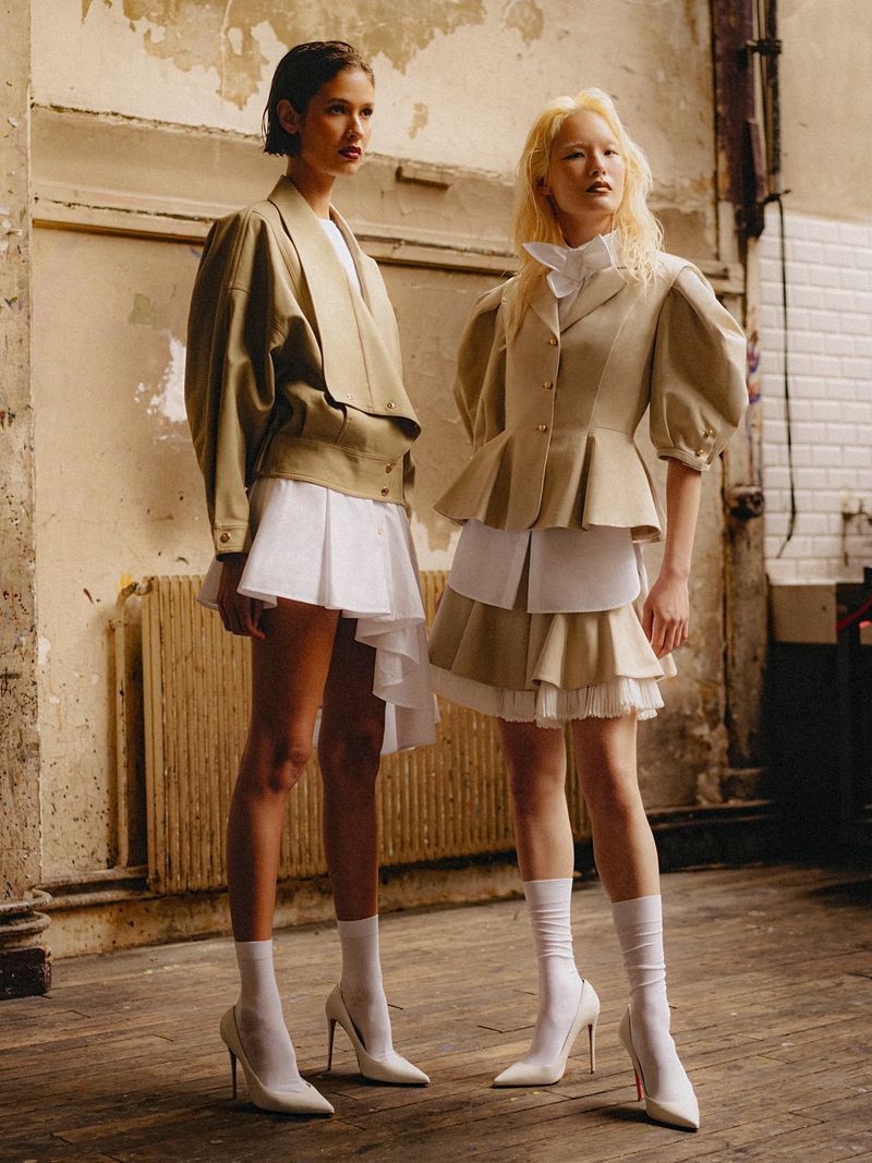 Two women in fashionable outfits pose indoors. Both wear jackets, white skirts, socks, and white high heels, standing on a parquet floor.