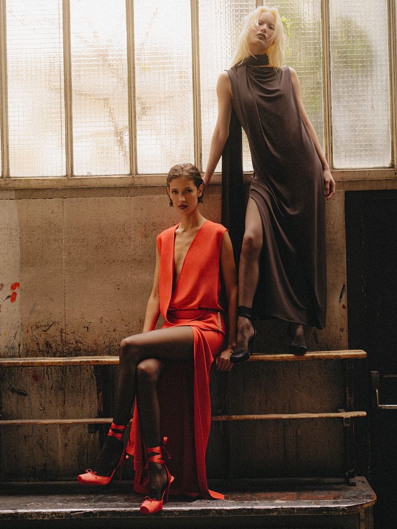 Two models in draped red and dark brown sleeveless evening dresses pose against large frosted industrial windows.