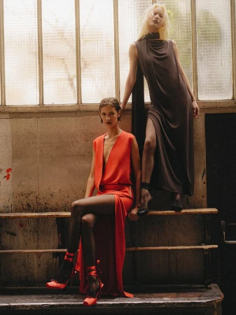 Two models in draped red and dark brown sleeveless evening dresses pose against large frosted industrial windows.