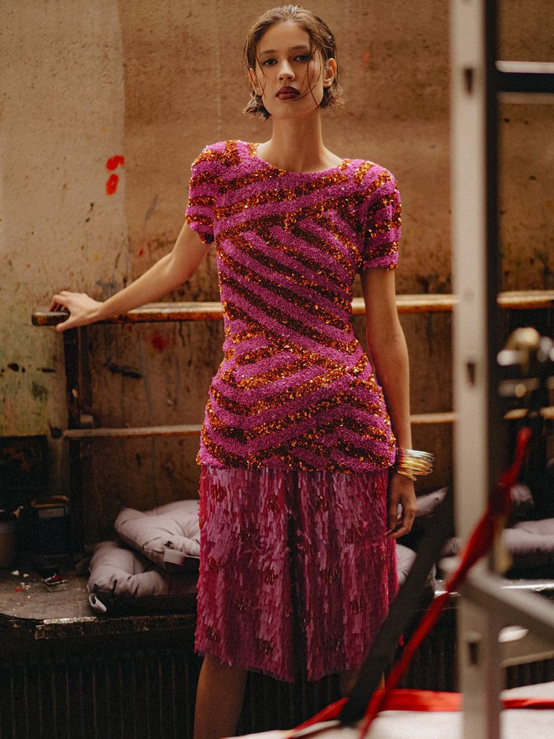 Model posing in a short-sleeve striped sequin top and magenta fringe skirt against a rustic studio backdrop.
