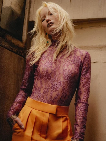 Bleach-blonde model in a sheer mauve floral lace top and bright orange trousers posing against a concrete wall.