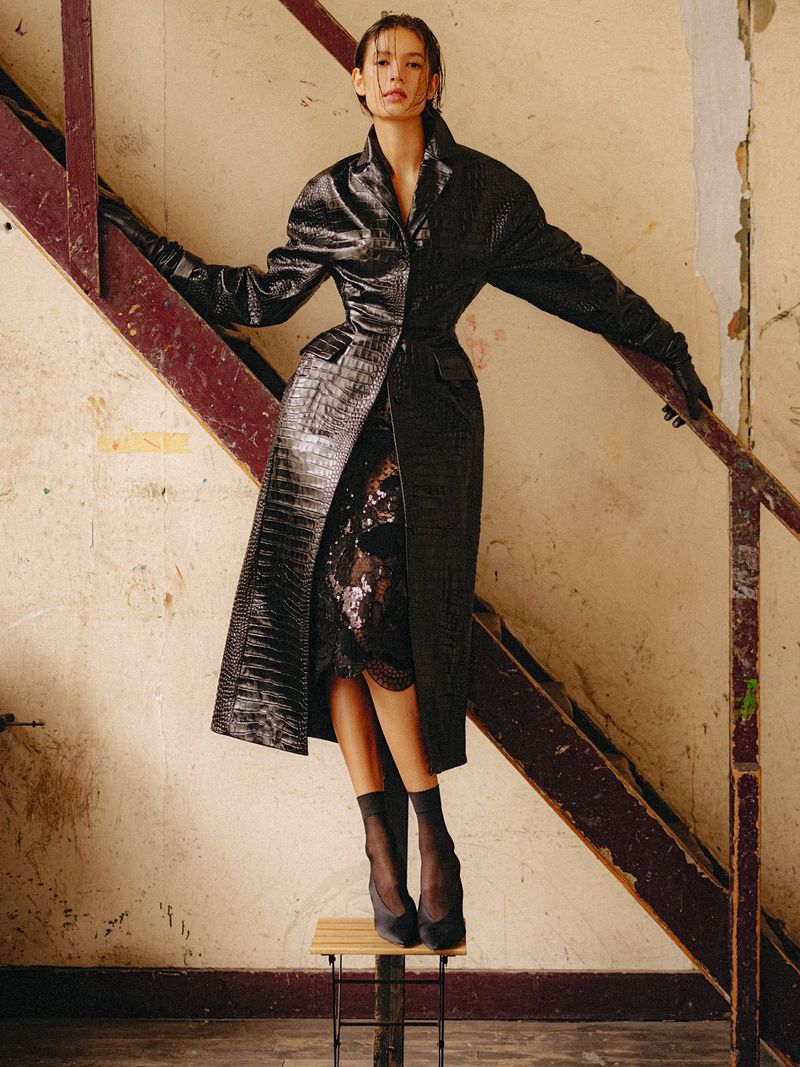 Model in a black textured coat and sheer black stockings standing on a stool beside a wooden staircase, shot by Ryan Tandya.