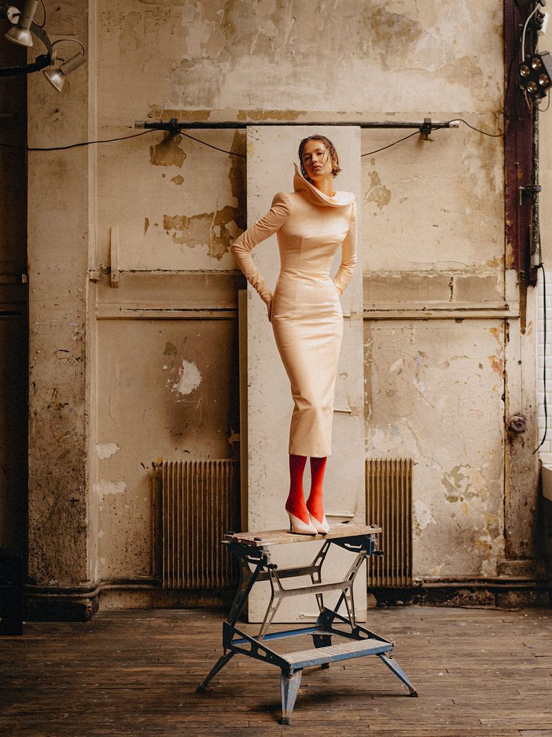 Model standing on a metal workbench in a weathered industrial interior wearing a structured beige dress and bright red tights.