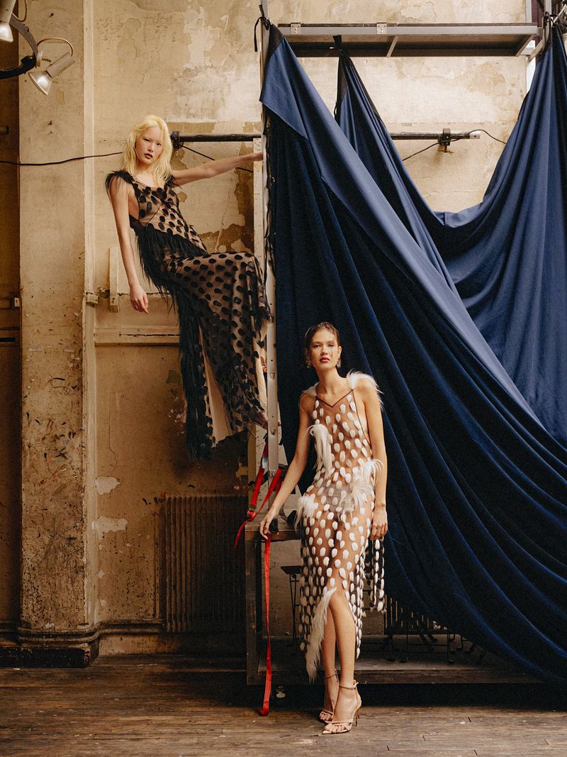 Two female models in sheer patterned dresses posing against industrial walls and large dark blue drapes.