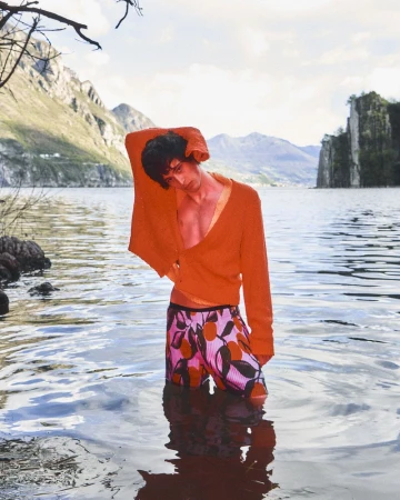 Model Federico Battisti stands waist-deep in a mountain lake wearing an orange knit cardigan and patterned swim shorts.