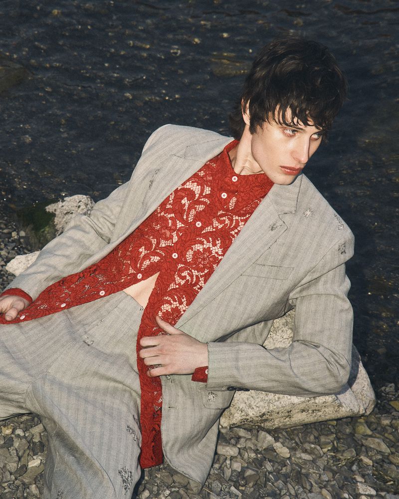 Model Federico Battisti reclining on a rocky shore wearing a grey pinstripe suit and an openwork red lace shirt.