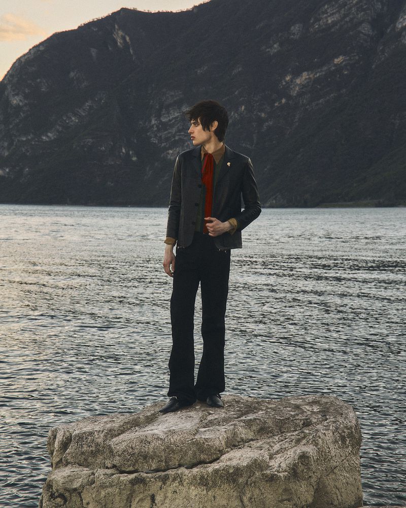 Federico Battisti stands atop a rock by a lake with mountains in the background wearing a dark tailored jacket and trousers.