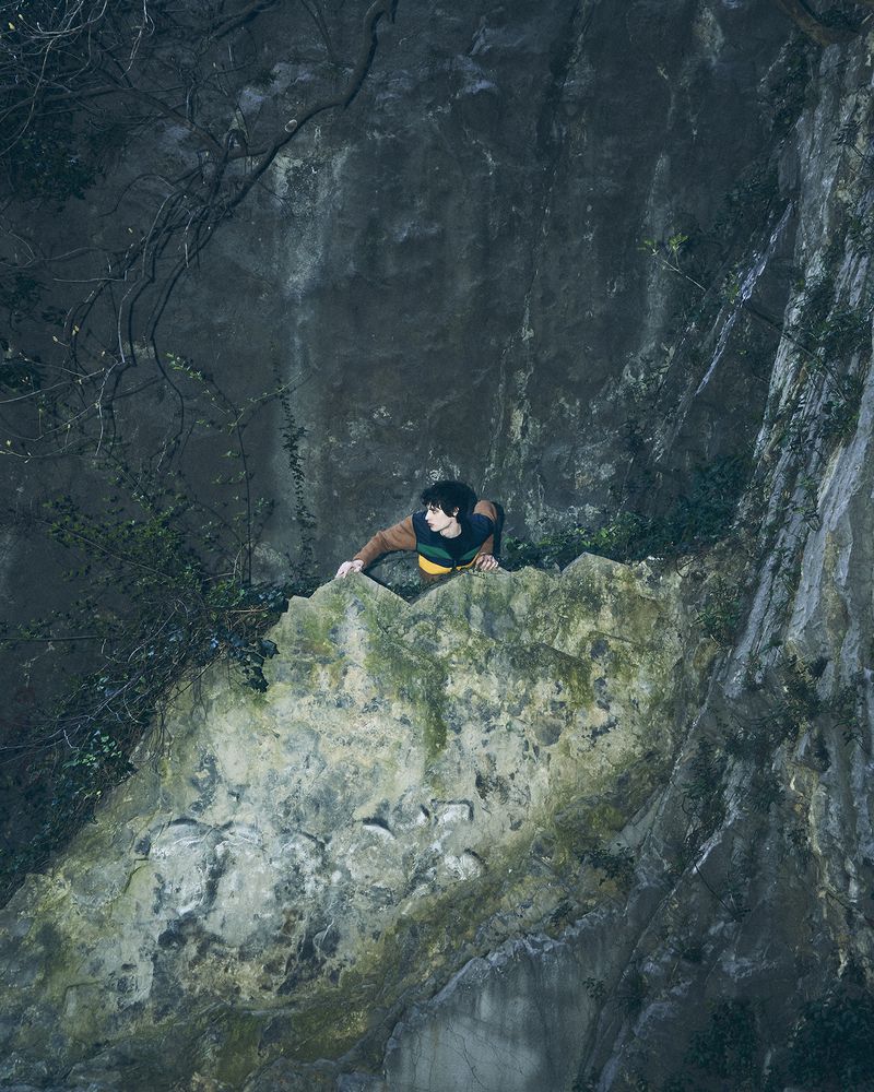 Model Federico Battisti climbs a mossy rock cliff face wearing a color-blocked brown and yellow sweater in an outdoor setting.