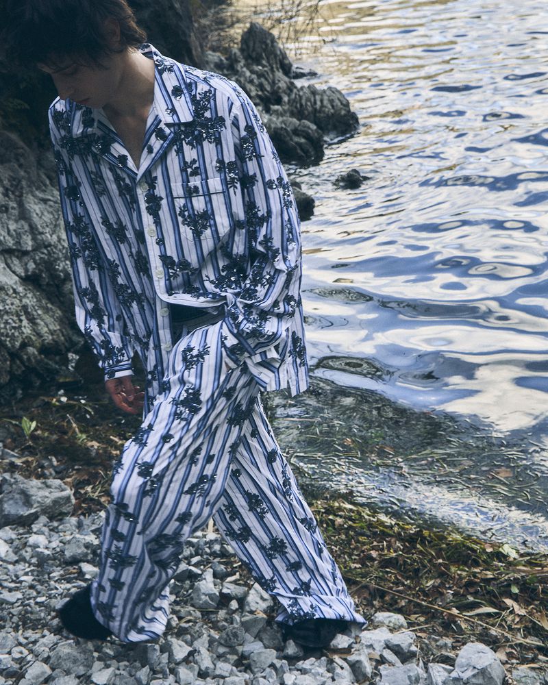 Model Federico Battisti walks along a rocky lakeshore wearing a blue and white striped floral matching set in an editorial shoot.