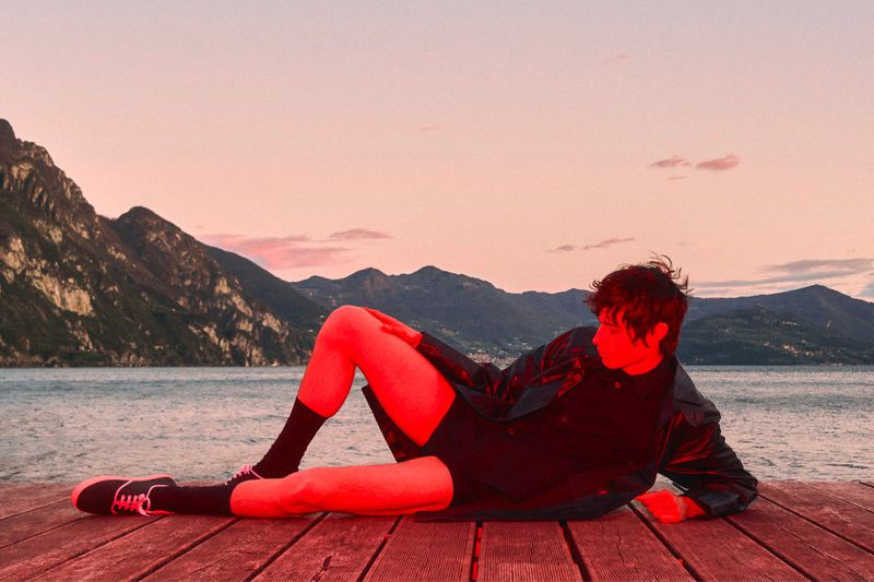 Model Federico Battisti reclining on a wooden dock overlooking a lake with mountains, illuminated by a vivid red light under a sunset sky.