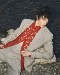 Model Federico Battisti reclining on a rocky shore wearing a grey pinstripe suit and an openwork red lace shirt.