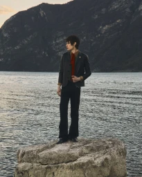 Federico Battisti stands atop a rock by a lake with mountains in the background wearing a dark tailored jacket and trousers.