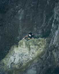 Model Federico Battisti climbs a mossy rock cliff face wearing a color-blocked brown and yellow sweater in an outdoor setting.