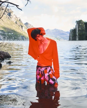 Model Federico Battisti stands waist-deep in a mountain lake wearing an orange knit cardigan and patterned swim shorts.