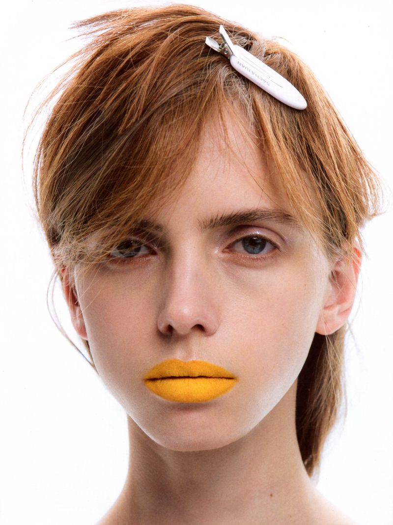 Close-up beauty portrait of a model with short auburn hair, a white styling clip, and bold matte yellow lipstick against a white backdrop.