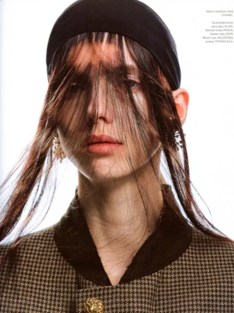 Close-up portrait of a model wearing a black headband and a patterned houndstooth jacket with hair covering eyes against a white background.