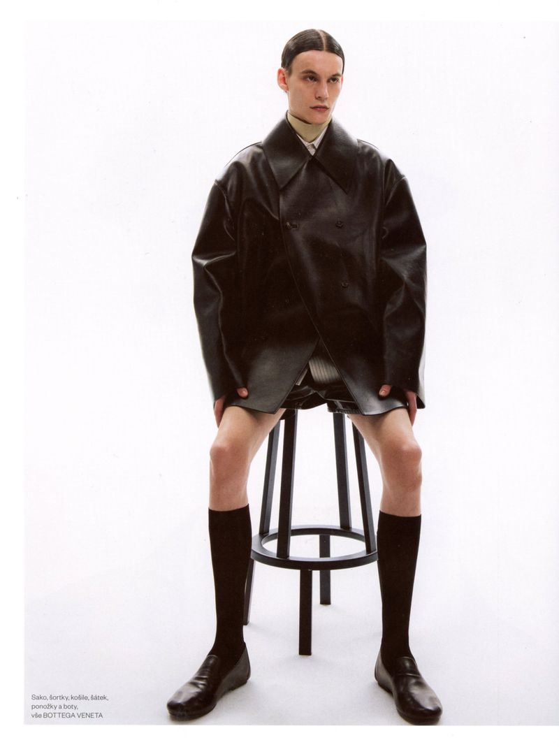 Model sitting on a black stool wearing an oversized black leather jacket and knee-high socks against a white studio background.