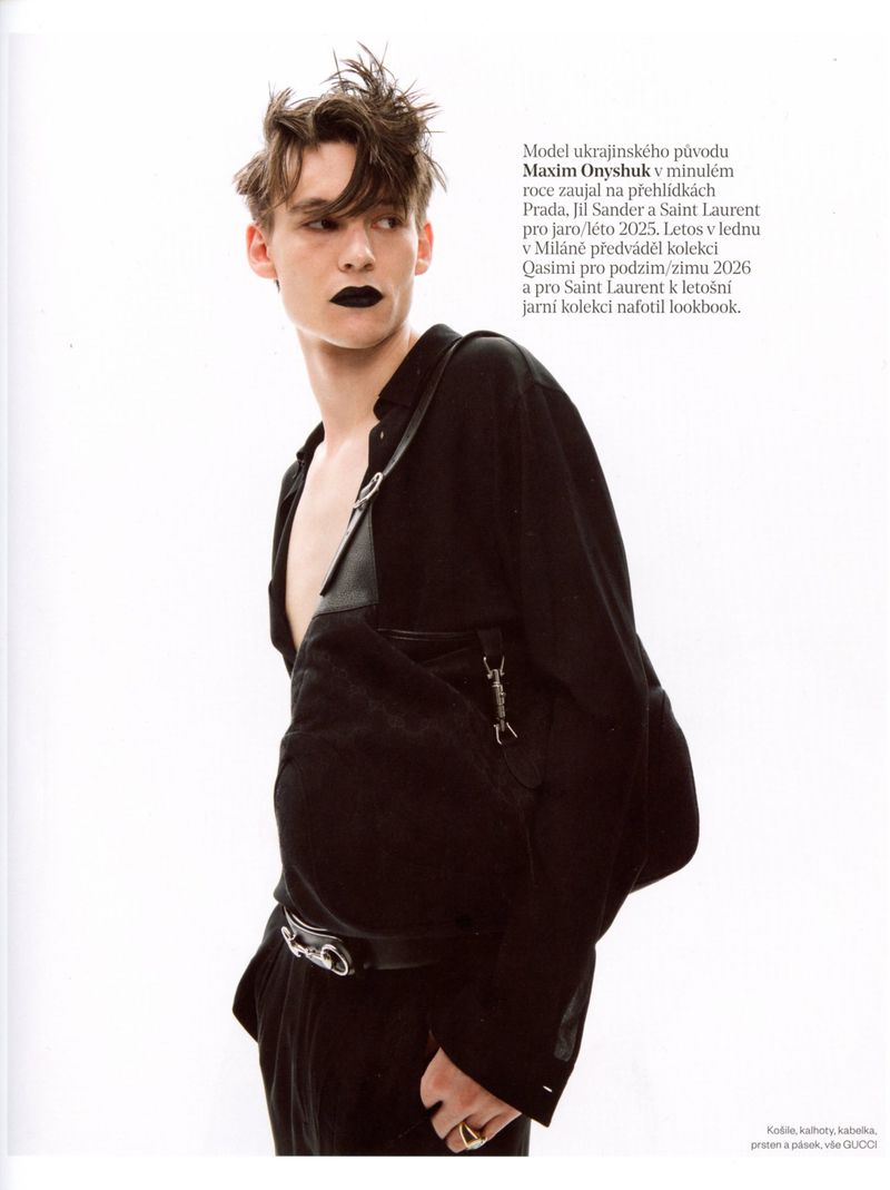 Male model with dark lipstick and textured hair wearing an open black shirt and crossbody strap against a white background.