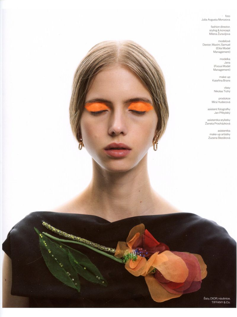 Model with closed eyes and vibrant orange eyeshadow wearing a black top with an orange fabric flower against a white background.