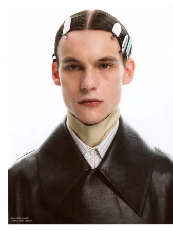 Male model with hair clips wearing a black leather coat and high collar scarf against a white studio background.