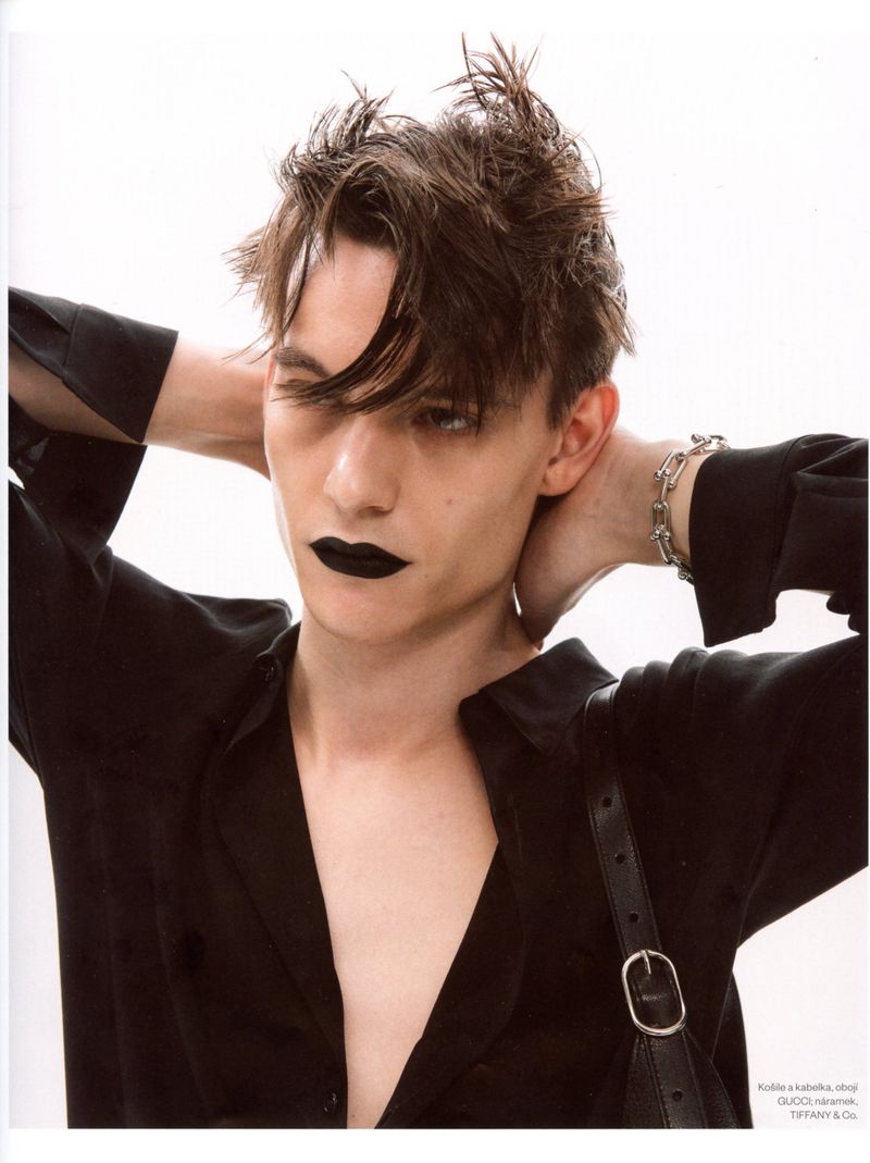 A young man with messy dark hair and black lipstick posing with hands behind his head in an unbuttoned black shirt on a white background.