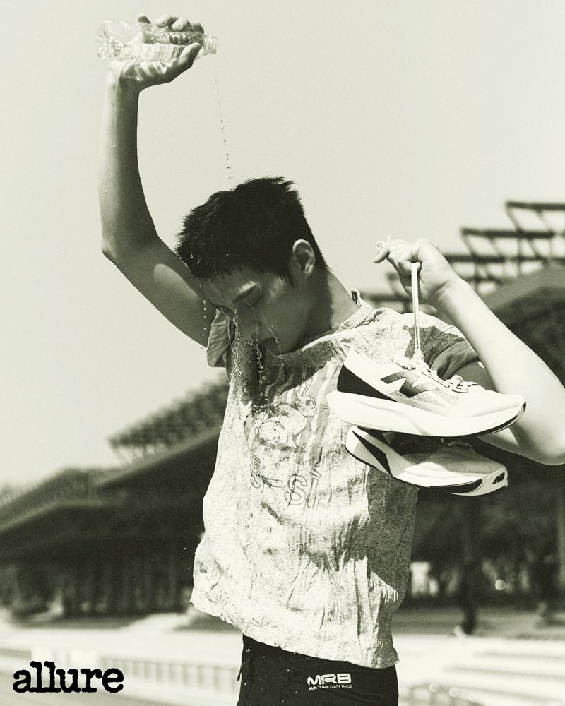Monochrome portrait of a sweaty man in activewear pouring water on his face outdoors, holding running shoes, with the Allure logo.
